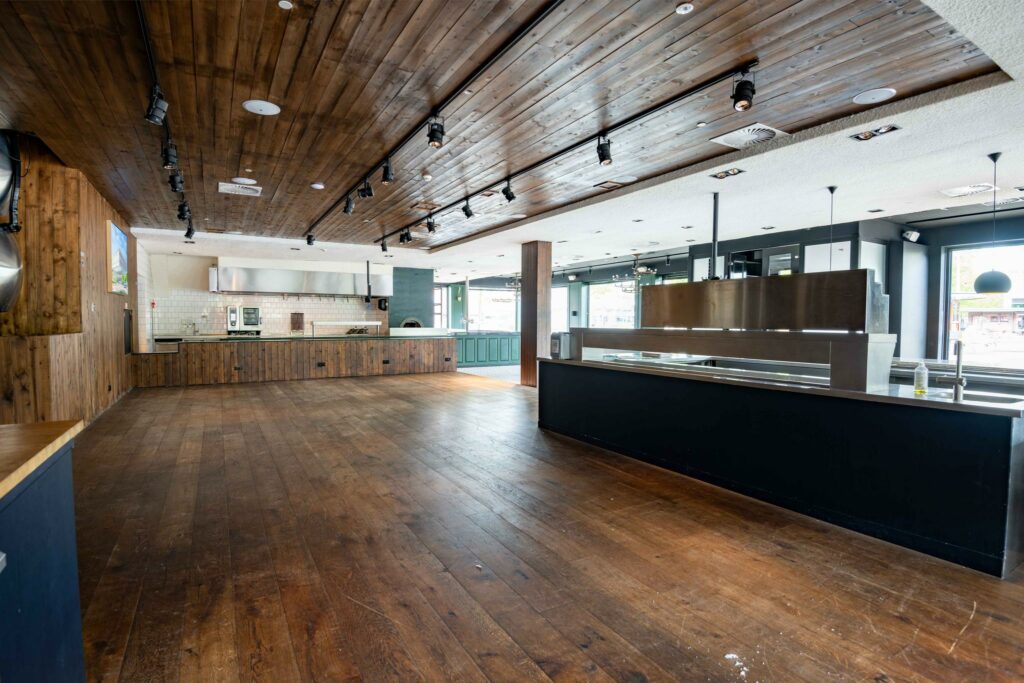 Ruim, modern ingericht horecagedeelte aan het Stadionplein met houten vloer en plafond, open keuken en grote ramen.