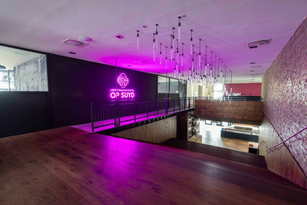 Moderne evenementenlocatie met houten vloeren, bakstenen muur en een felroze neonbord met de tekst "Meetings & Events Op Suyd".