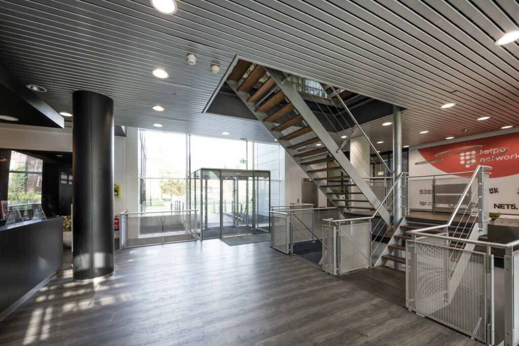 Moderne entreehal van een kantoorpand aan het Rietlandpark met glazen schuifdeuren, houten trap en metalen leuningen.