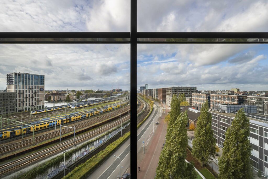 Uitzicht op station Amsterdam Rietlandpark met treinsporen, een weg en omliggende moderne gebouwen.