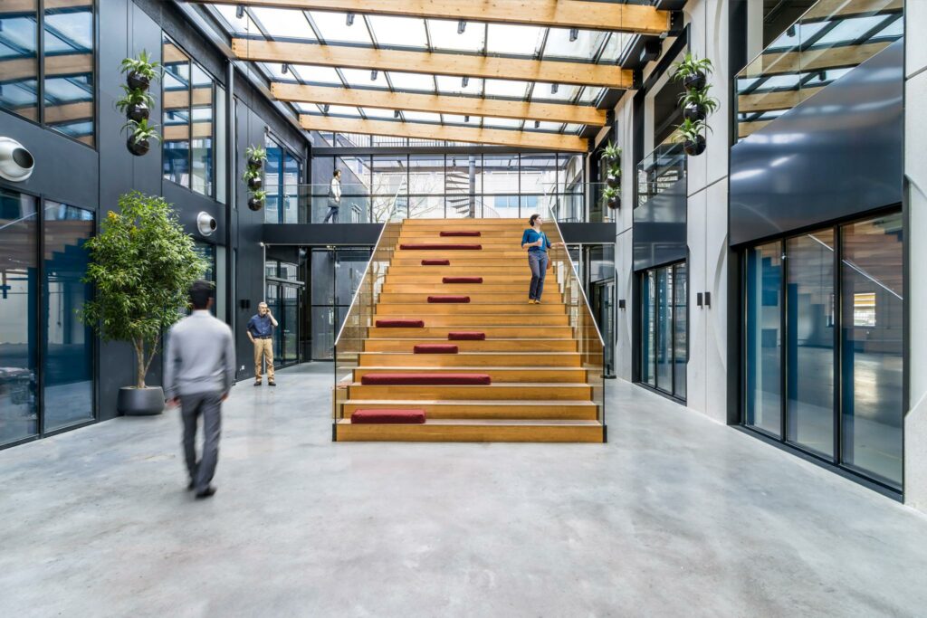 Moderne bedrijfsruimte aan de Laarderhoogtweg met grote houten tribunetrap, glazen dak en personen in gesprek en beweging.