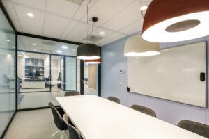 Vergaderruimte met glazen wanden, een lang wit tafeltje, grijze stoelen, een whiteboard en hanglampen aan het plafond in een modern kantoor aan de Paalbergweg.