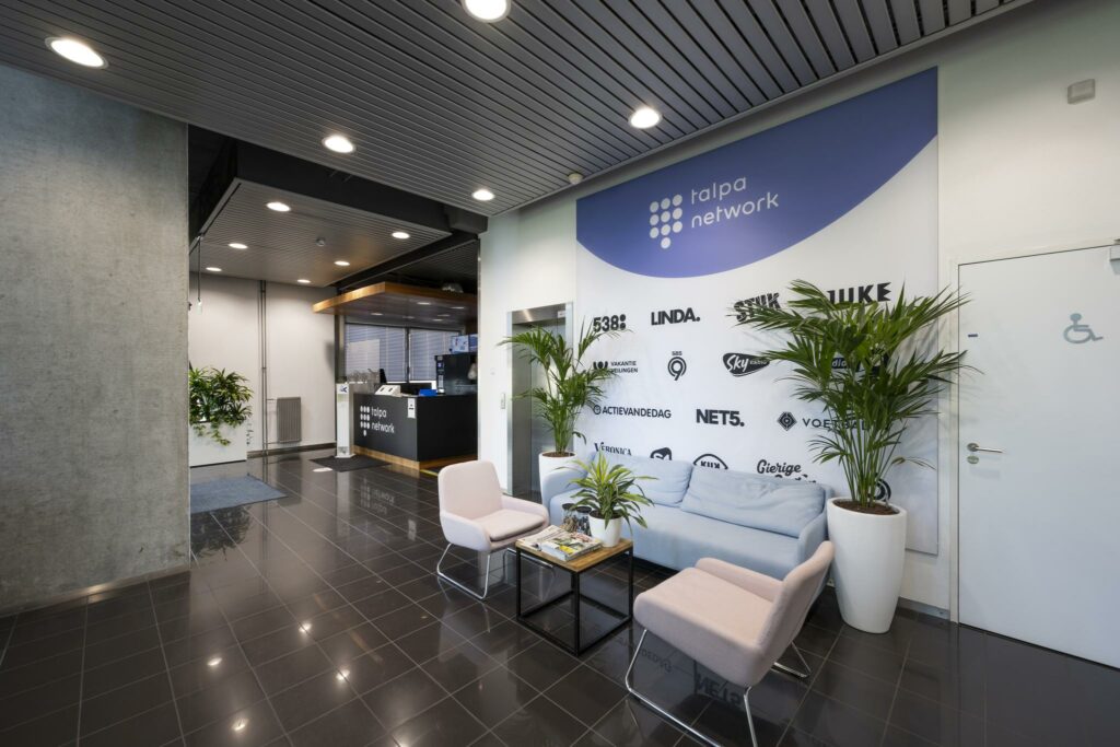 Moderne wachtruimte bij Talpa Network met zitgelegenheid, planten en een muur met logo’s van mediakanalen.