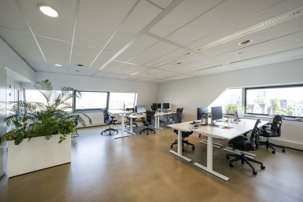 Modern kantoorinterieur in Rietlandpark met verstelbare bureaus, bureaustoelen, computers en grote ramen met stadszicht.
