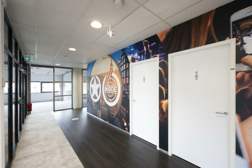 Moderne gang met witte toiletten en een kleurrijke muurfoto met het logo van Rietlandpark 6 Inside.