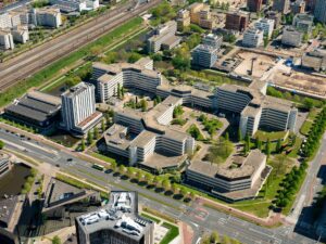 Luchtfoto van het kantoorgebouwcomplex aan de Hoogoorddreef in Amsterdam-Zuidoost, omringd door wegen, spoorlijnen en andere gebouwen.