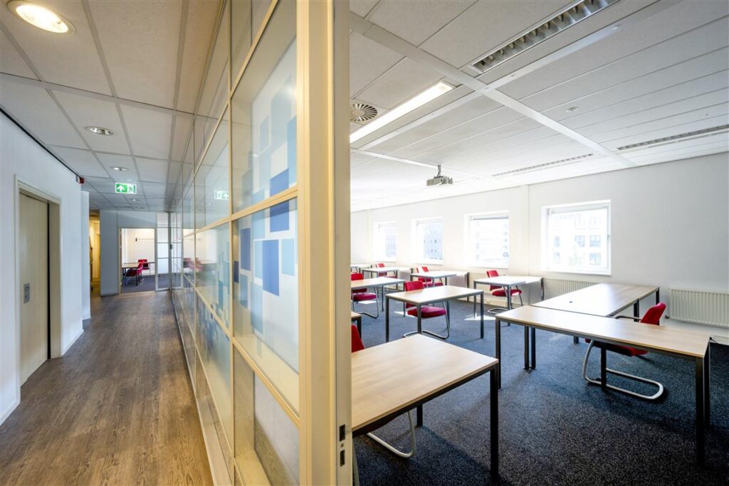 Lichte en moderne les- of vergaderruimte aan de Paasheuvelweg met houten tafels en rode stoelen.