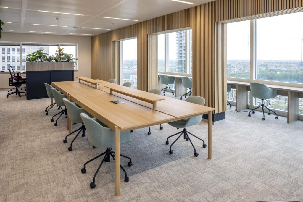 Modern open-plan office space at Amstelplein with shared wooden desks, ergonomic chairs, and large windows offering a city view.