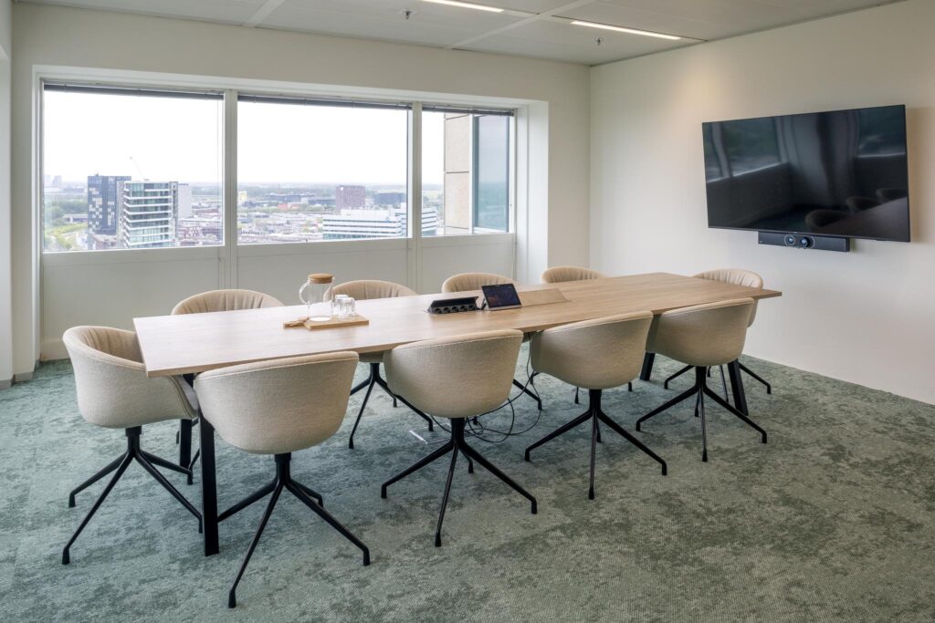 Modern meeting room at Amstelplein with a long wooden table, eight chairs, a large screen, and a view over the city.