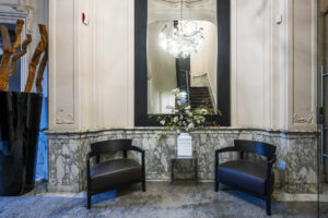 Stijlvolle entree aan de Herengracht met marmeren lambrisering, twee zwarte fauteuils, een witte vaas met bloemen en een grote spiegel waarin een trap wordt weerspiegeld.