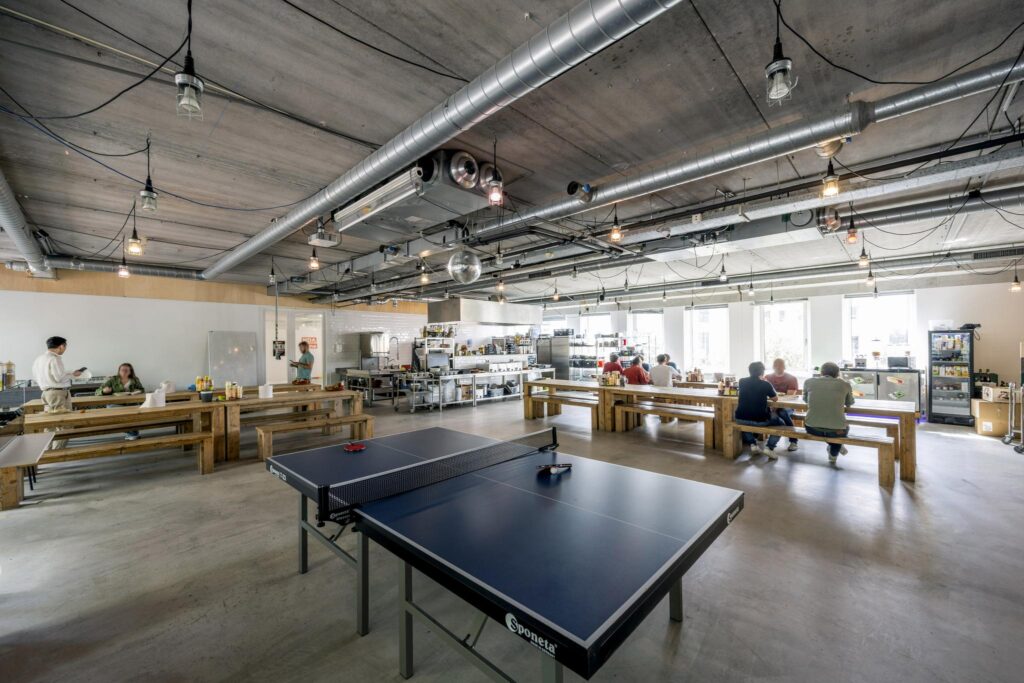 Moderne bedrijfskeuken met lange houten tafels, een tafeltennistafel en mensen die eten of praten in een open en industriële ruimte aan de Naritaweg.