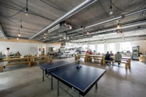 Moderne bedrijfskeuken met lange houten tafels, een tafeltennistafel en mensen die eten of praten in een open en industriële ruimte aan de Naritaweg.
