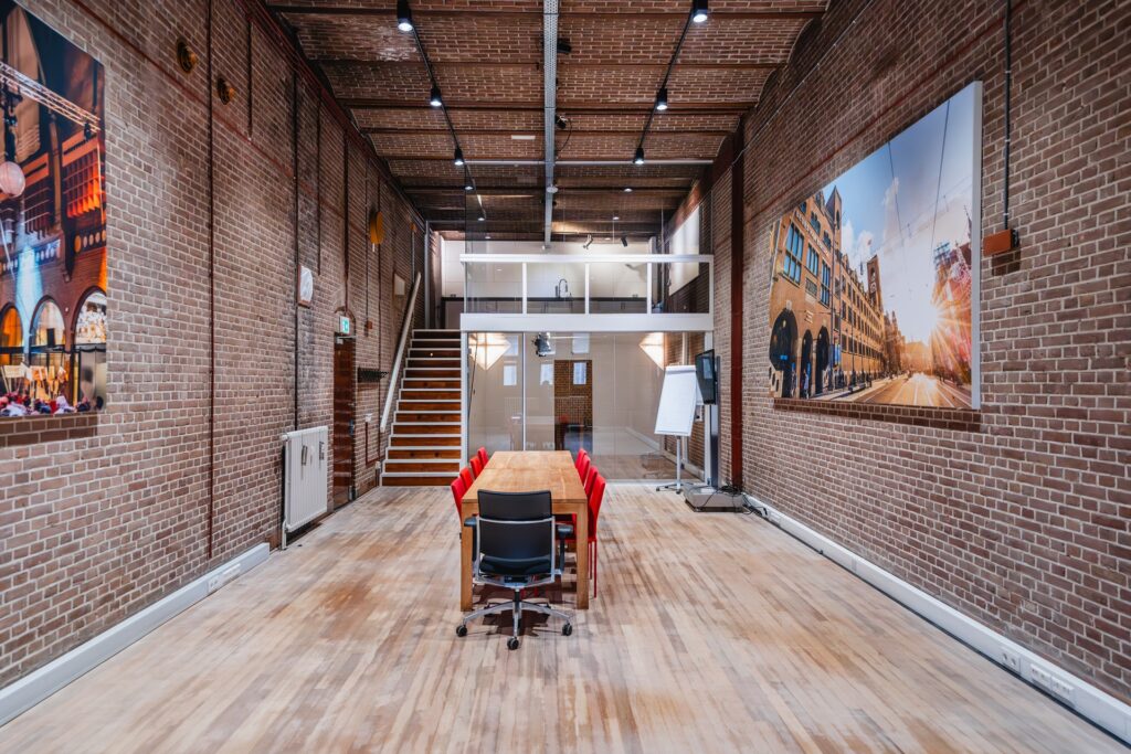 Modern meeting room with brick walls, wooden floor, red chairs, and a large photo of Damrak street in Amsterdam.
