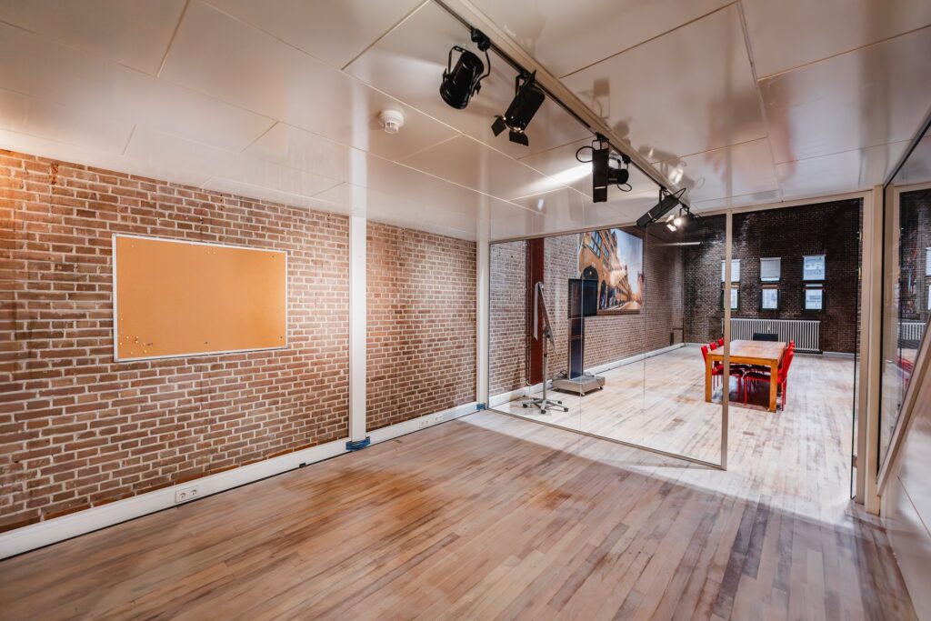 Modern meeting room with exposed brick walls, a wooden floor, glass partitions, and a table with red chairs.