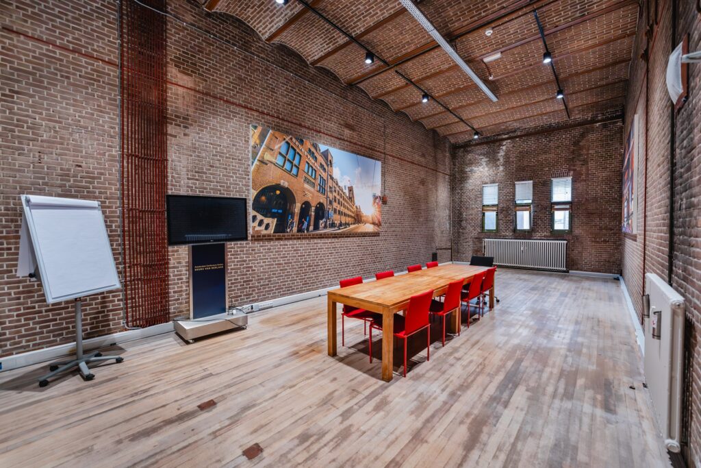 Modern meeting room at Damrak with a long wooden table, red chairs, and exposed brick walls.