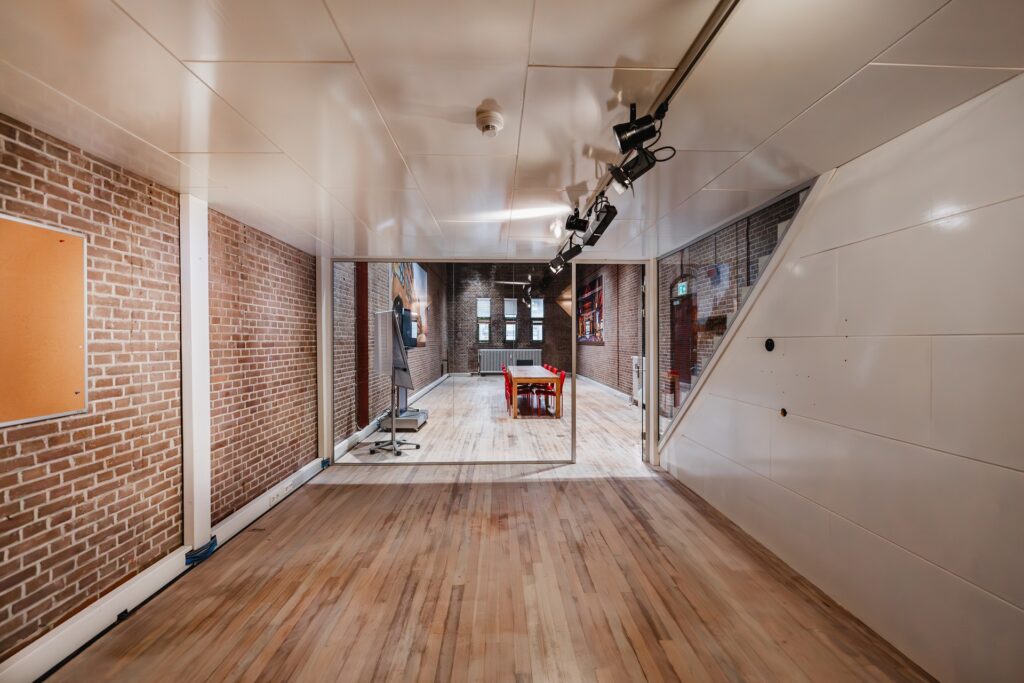 Modern meeting room at Damrak 173 with exposed brick walls, wooden flooring, and red chairs around a central table.