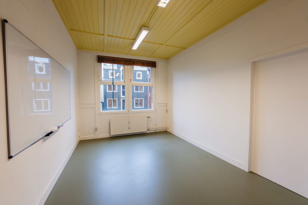 Small empty classroom at Damrak with a whiteboard, yellow ceiling, and large window.