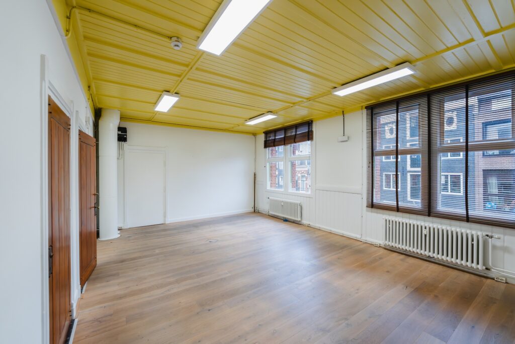 Bright empty room with yellow ceiling, wooden floor, and large windows overlooking Damrak.