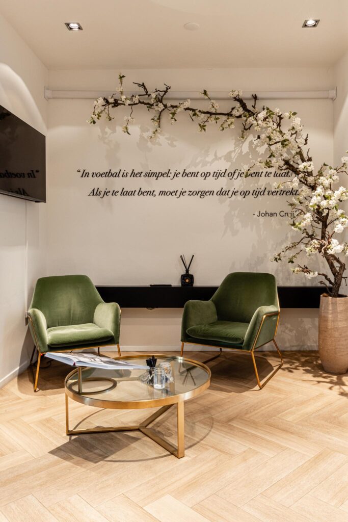 Stijlvolle wachtruimte met twee groene fauteuils, een ronde glazen salontafel en een muurquote van Johan Cruijff.