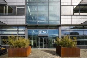 Hoofdentree van het kantoorgebouw GO WEST aan de Kabelweg 47 in Amsterdam, met grote glazen gevel en plantenbakken.