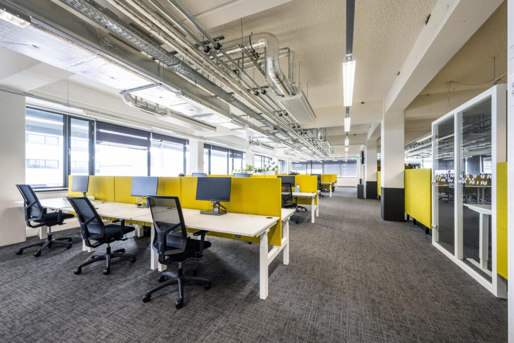 Modern en licht kantoorinterieur aan de Kabelweg met open werkplekken en gele scheidingspanelen.