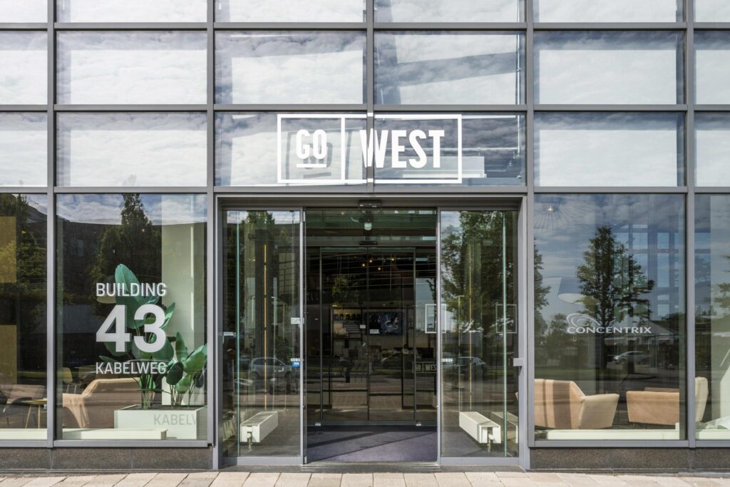 Glazen entree van kantoorgebouw Go West aan de Kabelweg 43 met zicht op de lobby en bedrijfsnamen.