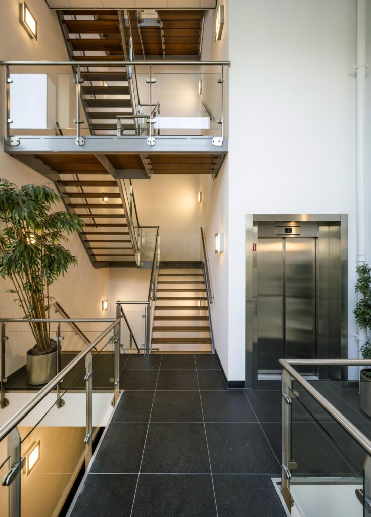 Moderne trappenhal in een bedrijfsverzamelgebouw aan de Sijsjesbergweg met glazen balustrades, een lift en planten.