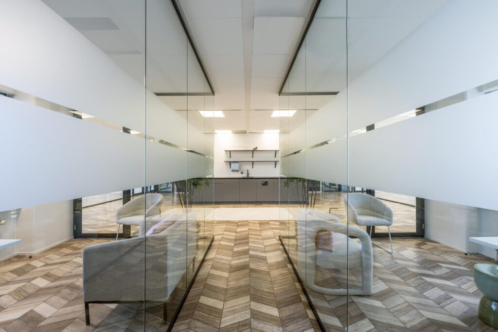 Modern office kitchenette with glass walls, wooden herringbone floor, and minimalist seating area on Overschiestraat.