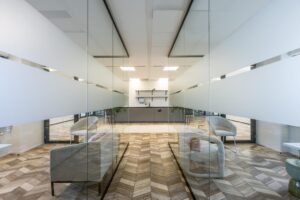 Modern office kitchenette with glass walls, wooden herringbone floor, and minimalist seating area on Overschiestraat.