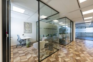 Modern office space at Overschiestraat with glass walls, wooden herringbone floor, and minimalist furnishings.