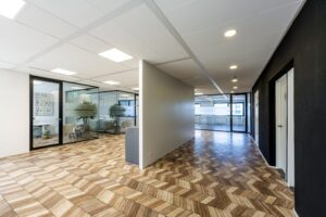 Modern office hallway on Overschiestraat with glass-walled meeting rooms and a herringbone wooden floor pattern.