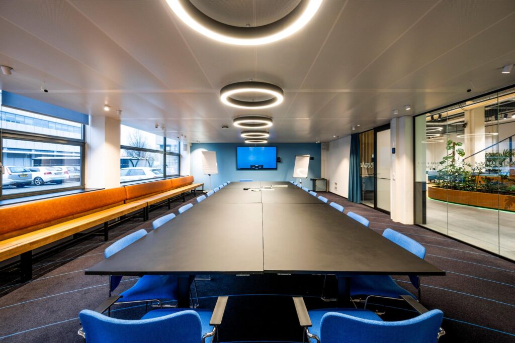 Moderne vergaderruimte aan de Kabelweg met lange conferentietafel, blauwe stoelen, presentatievoorzieningen en grote ramen.