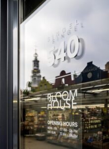 Winkelraam met opschrift "Singel 540" en "Bloom House" met openingstijden, met weerspiegeling van de bloemenmarkt en de Munttoren in Amsterdam.