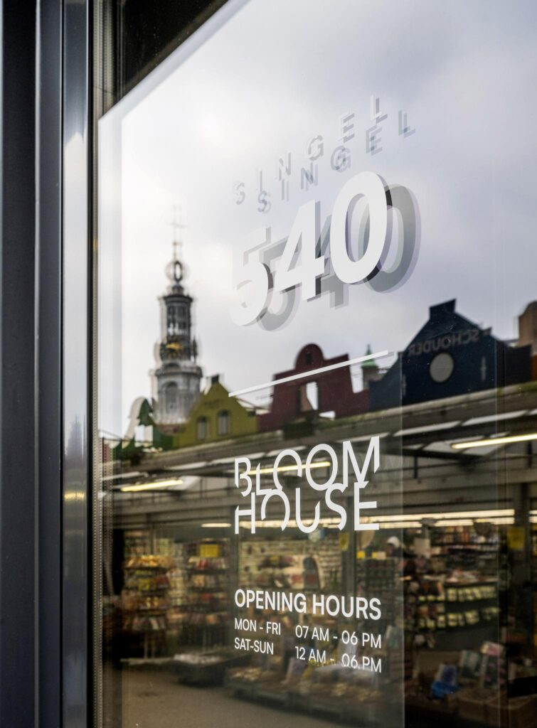 Winkelraam met opschrift "Singel 540" en "Bloom House" met openingstijden, met weerspiegeling van de bloemenmarkt en de Munttoren in Amsterdam.