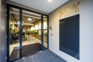 Ingang van het Bloom House-gebouw met glazen deuren en een modern interieur vol planten.