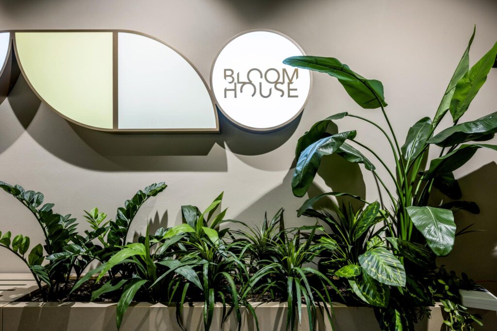Wanddecoratie met het verlichte logo van Bloom House boven een plantenbak met diverse tropische kamerplanten.