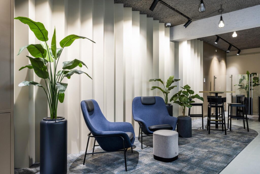 Moderne wachtruimte met blauwe loungefauteuils, hoge planten en een stijlvolle lichtinval tegen een geribbelde wand.