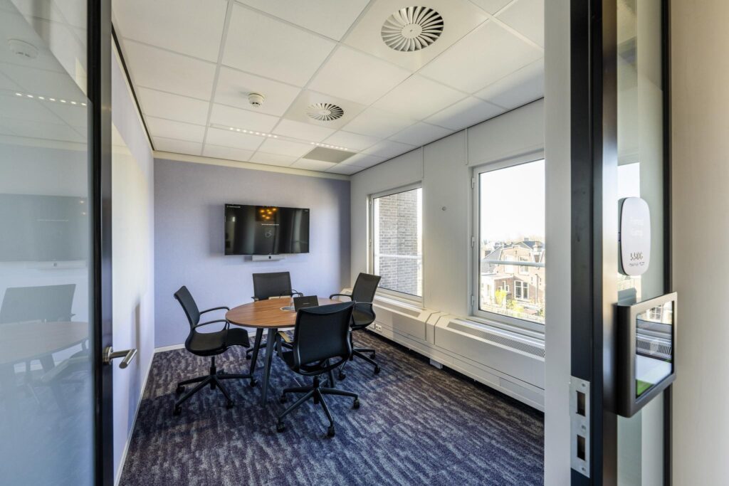 Modern meeting room at Vijzelgracht with a round table, four chairs, a wall-mounted screen, and large windows offering a city view.