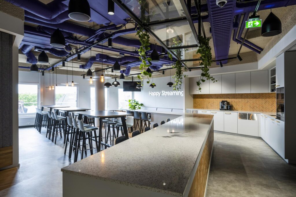 Modern office kitchen and dining area with high tables, hanging plants, and a "Happy Streaming" neon sign.