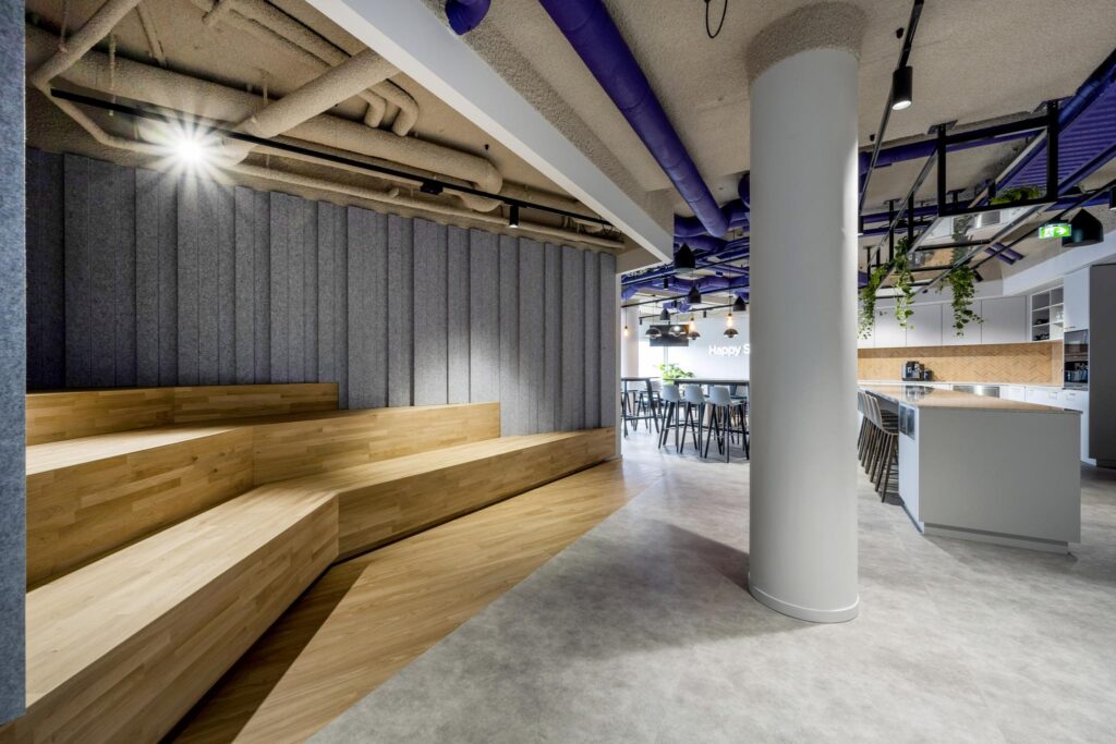 Modern office interior with wooden bleacher seating, an open kitchen area, and contemporary industrial design elements.