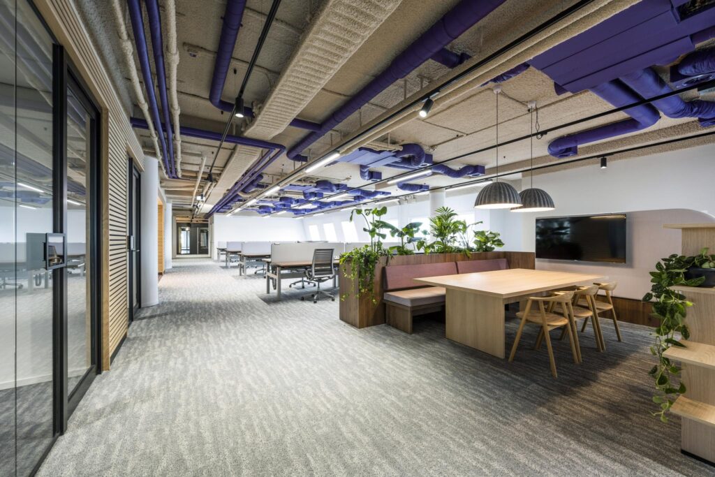 Modern open-plan office space on Vijzelgracht featuring shared desks, a wooden meeting table with built-in seating, and exposed purple ceiling pipes.