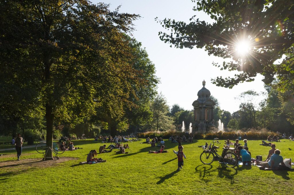 People relaxing and enjoying the sun on the grass near a historic fountain in Vondelpark, Amsterdam.