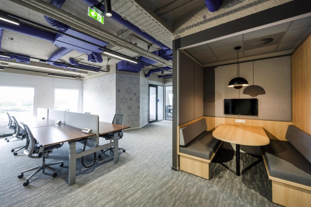 Modern open office space with shared desks and a built-in meeting nook featuring a screen and pendant light.