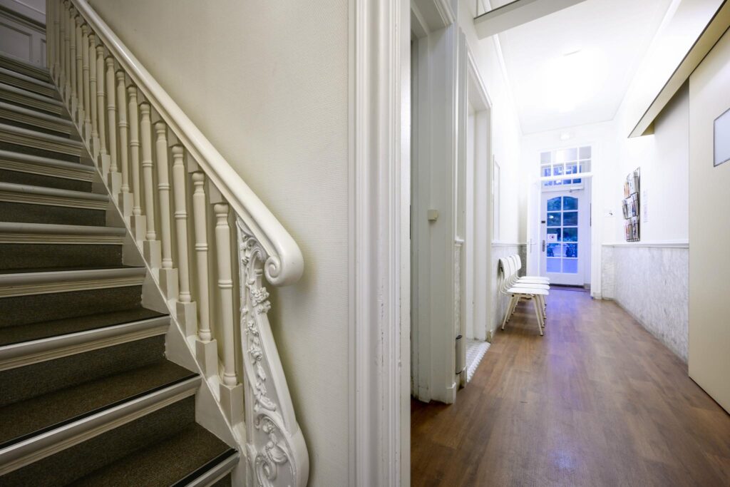 Bright hallway with wooden floor, white chairs, and a staircase featuring ornate banisters at Concertgebouwplein.