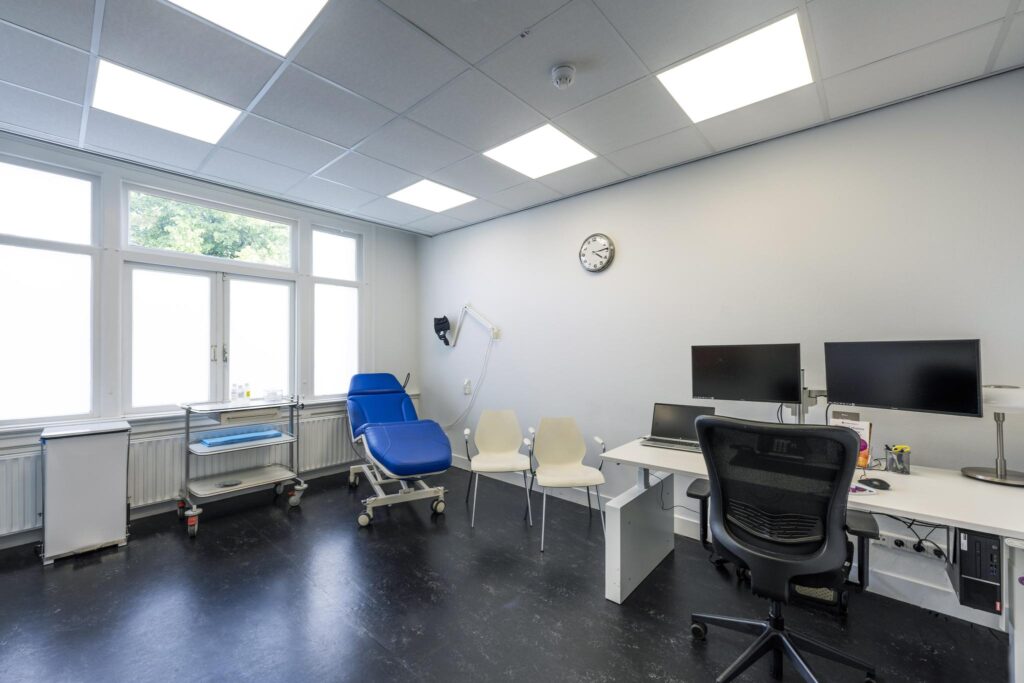 Modern medical consultation room with a blue examination chair, desk setup, and medical equipment.