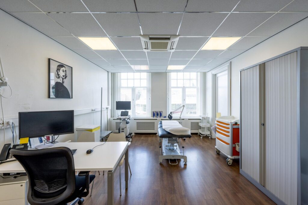 Bright and modern medical examination room with a treatment table, desk, medical equipment, and large windows.