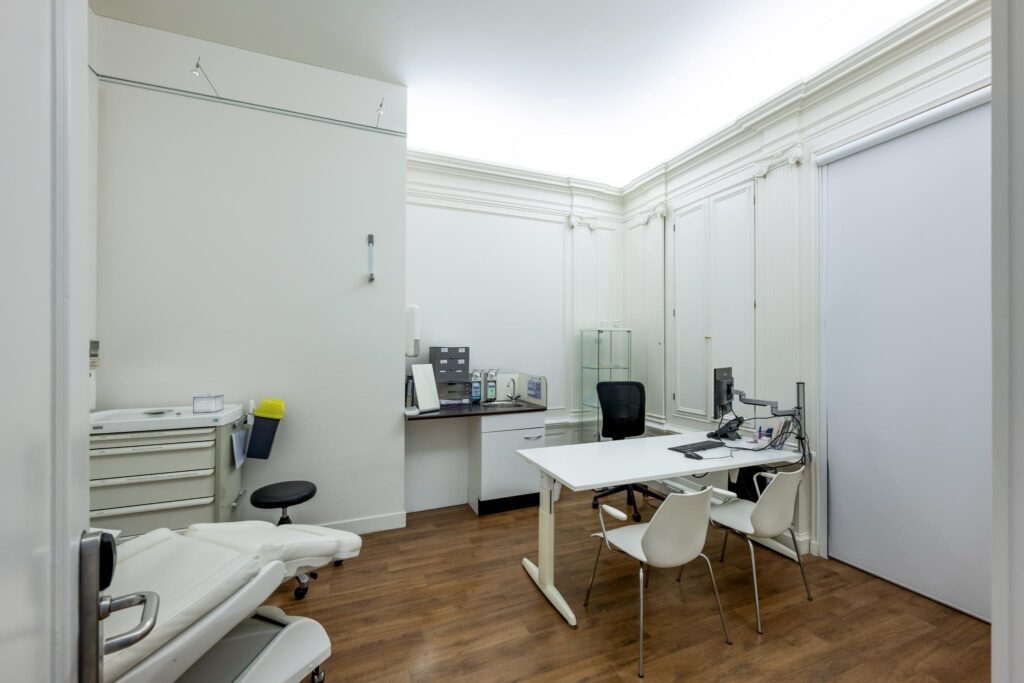 Modern medical consultation room with a desk, examination chair, and computer equipment.