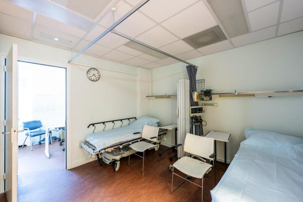 Hospital room with two beds, medical equipment, and chairs, viewed from an open door.
