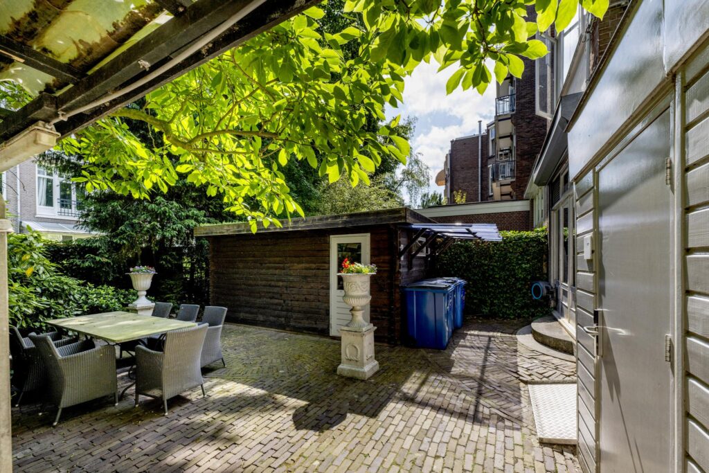 Paved backyard area with outdoor dining set, greenery, a wooden shed, and recycling bins at Concertgebouwplein.