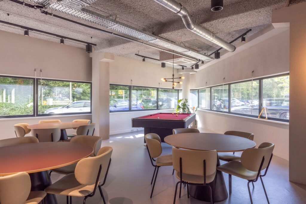 Modern common area with round tables, chairs, and a red pool table near large windows.